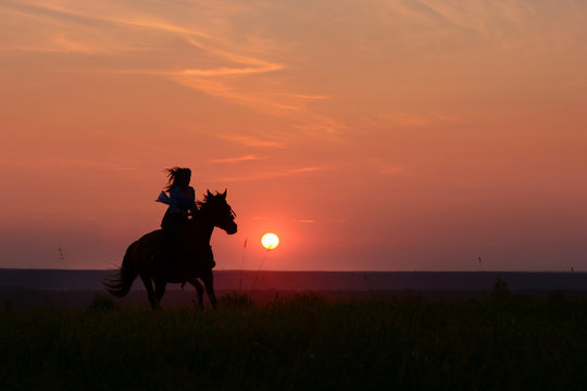 Galloping horse on sunset. Horseback woman riding gallop with red rising sun on horizon. Horse hiking on beautiful colorful background with equine and girls silhouette. 