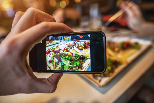 Close up focus view of hand and mobile while taking a photo of food on the plate in a restaurant for social media.