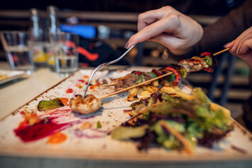 Close up focus view of hand and fork while taking meat from fried skewers on a wooden stick.