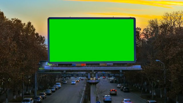 A billboard with green chroma key on the background of fast moving cars at sunset. Time Lapse video. The camera is approaching