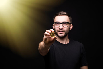 Young handsome man in glasses in black shirt pointed golden bitcoin on camera isolated on black background. Tonned image
