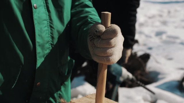 Close up hand in worker gloves hammering two wood bars to hole in wooden block outside in winter, snow on background
