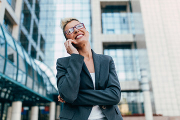 Young happy business woman standing in front of big modern building. She smiling and talking on her cell phone.