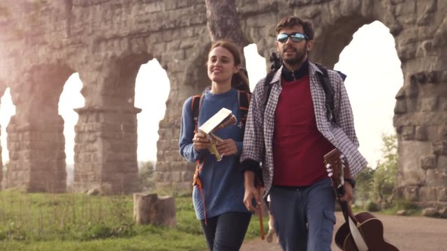 Young couple backpackers tourists walking with map guide near roman aqueduct arches in parco degli acquedotti park ruins in rome at sunrise with guitar and sleeping bag slow motion steadycam
