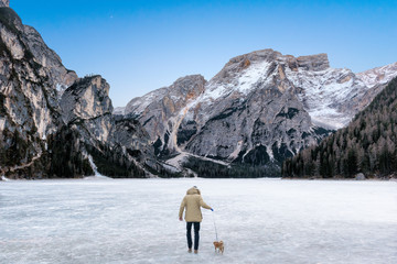 Man leads a dog on a leash for a walk on a frozen lake covered with snow surrounded by mountains. Braies Lake, Trentino, Iraly Clear sky