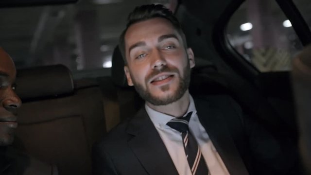 Handheld shot of cheerful bearded businessman in suit chatting with male African colleague while seating on backseat of moving car
