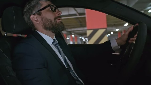 Shot from passenger seat of moving car of bearded businessman in glasses driving and looking around while searching for free space in underground parking lot