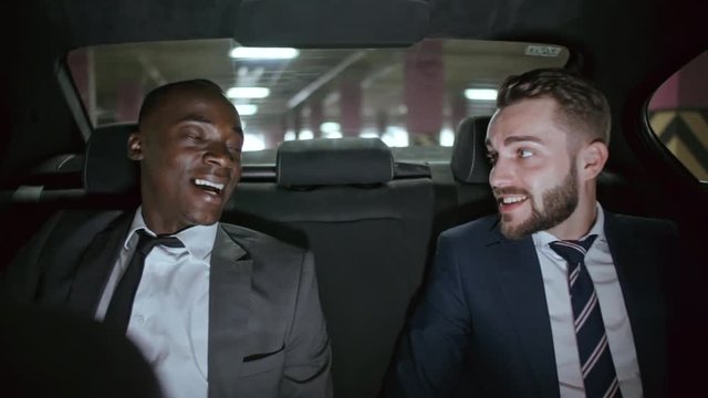 Cheerful African businessman in suit and his Caucasian colleague with beard sitting on backseat of moving car and chatting