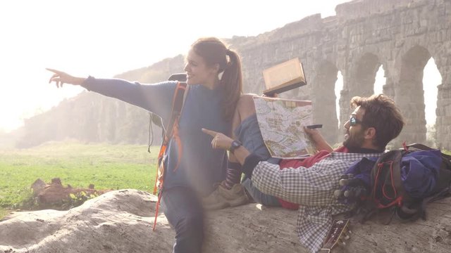 Happy young couple backpackers tourists sitting lying on a log trunk reading map guide pointing ancient roman aqueduct ruins in parco degli acquedotti park in rome at sunrise romantic with sleeping