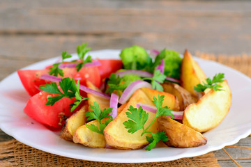Fried potatoes with vegetables. Fried potato wedges, cooked broccoli, fresh tomato slices, red onion, green parsley on a serving plate and on a wood background. Vegetarian main course recipe. Closeup