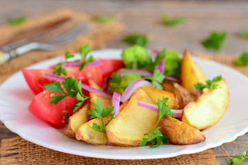 Roasted potatoes and vegetables. Roasted potato wedges, boiled broccoli, fresh tomato slices, red onion, green parsley on a serving plate and on a wooden background. Vegetarian entree recipe. Closeup
