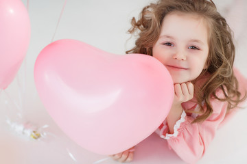 A little girl of 6 years with long curly hair, a beautiful smile, sits alone in a large bright room with lots of pink balloons in the shape of a heart. Valentine's Day and party celebration.