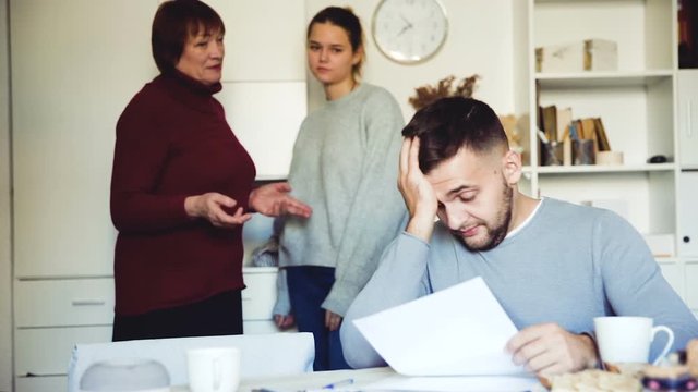 Upset guy sitting with documents while dissatisfied girl with older woman standing behind him