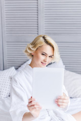 selective focus of smiling young woman in bathrobe using digital tablet in bedroom