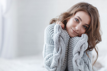 Obraz premium Cute young girl in a gray knitted sweater. Beautiful woman is relaxing in a white bedroom. Beautiful women in winter clothes are waking up in the morning. Woman wearing a sweater in a white bedroom.