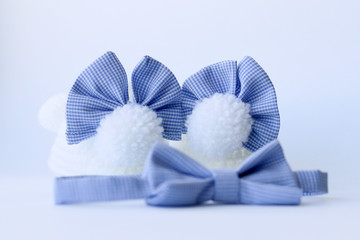 Cute knit white baby booties with fluffy bunny tails, plaid blue bows and little bow tie on white background. Focus on bows. Newborn concept. Close-up