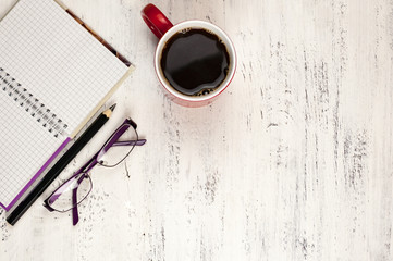 Pencil, notebook, glasses and a cup of coffee on a white wooden table.