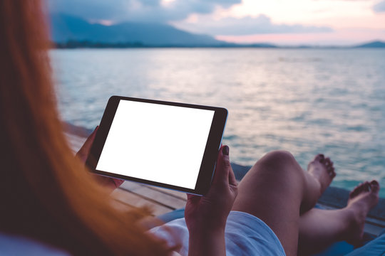 Mockup image of a woman using black tablet pc with blank white desktop screen while sitting by the sea with blue sky background