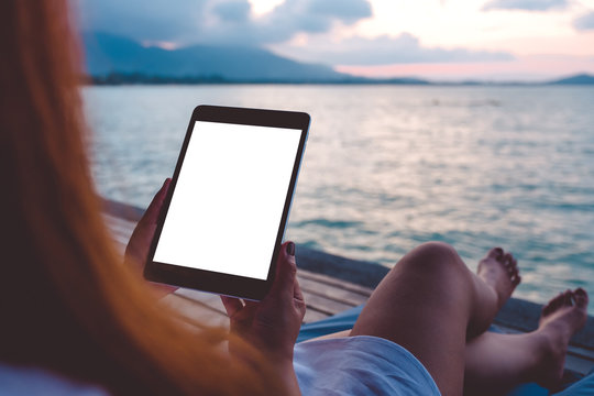 Mockup image of a woman using black tablet pc with blank white desktop screen while sitting by the sea with blue sky background
