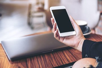 Mockup image of a woman's hand holding white mobile phone with blank black desktop screen and laptop on wooden table in cafe