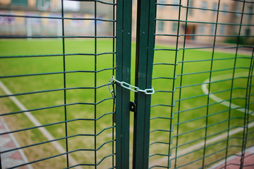 Close up metallic net-shaped green fence that closed and wrapped by chain on a background of school football field