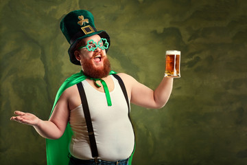 A fat man with a beard in St. Patrick's suit is smiling with a mug of beer on a green background.