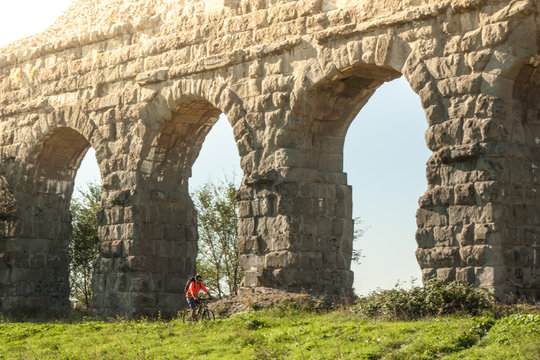 Cyclist riding bike on dirt road under ancient roman aqueduct in a park. Young attractive athletic man with orange sportswear and backpack in parco degli acquedotti in Rome