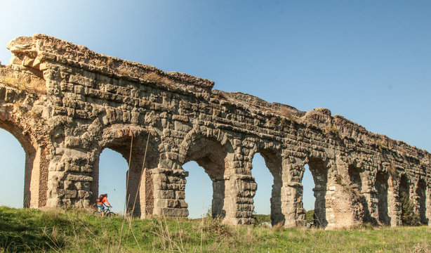 Cyclist riding bike on dirt road under ancient roman aqueduct in a park. Young attractive athletic man with orange sportswear and backpack in parco degli acquedotti in Rome