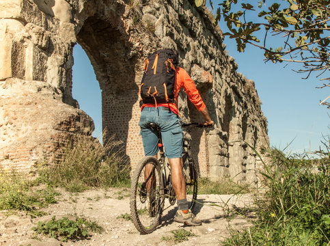 Cyclist riding bike on dirt road under ancient roman aqueduct in a park. Young attractive athletic man with orange sportswear and backpack in parco degli acquedotti in Rome
