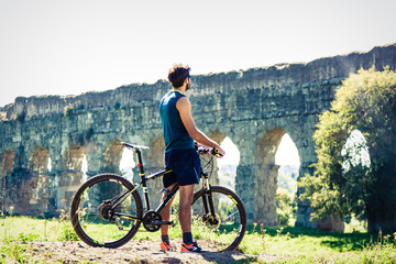 Cyclist standing on bike on dirt road in front of ancient roman aqueduct. Young attractive athletic man with blue t-shirt and shorts sportswear in acquedotti park in rome