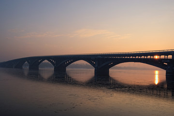 Picturesque view over the Metro (Subway) Bridge over the Dnipro river in Kyiv, Ukraine. Sunrise at winter morning. The red disk of the sun peeps out from behind the bridge