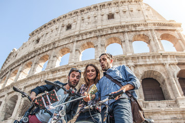 Three happy young friends tourists with bikes and backpacks at Colosseum in Rome taking pictures with smartphone and selfie stick having fun. Ground shot with lens flare.