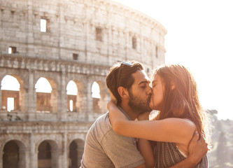 Romantic happy couple young tourists in love kissing cuddling in front of colosseum in rome at sunset with lens flare.