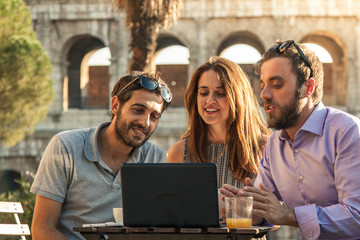 Three happy friends using laptop having video call sitting at bar restaurant table in front of colosseum in rome at sunset.