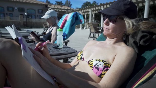 Closeup of pretty woman in sunglasses, hat, and bikini outside by a pool reading a book and highlighting in the book.