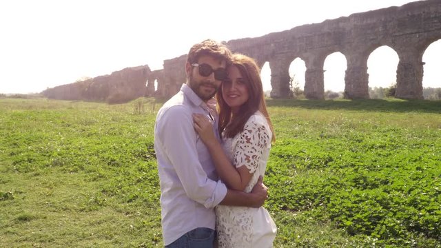 Come with me. Young beautiful girl with hat hugs man boyfriend posing for photo in front of roman aqueduct arches in parco degli acquedotti park ruins in rome romantic lovely couple slow motion