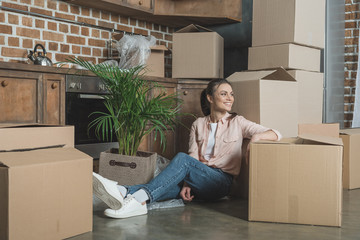 Fototapeta na wymiar happy young woman smiling and looking away while sitting with cardboard boxes in new apartment