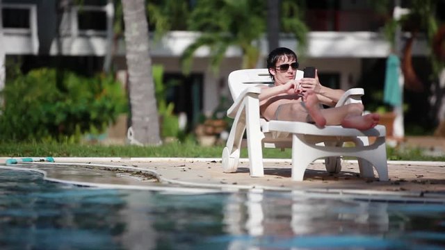 Young man wearing sunglasses uses mobile phone while lying on sunbed by swimming infinity pool background. 1920x1080, hd