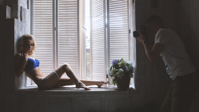 Young blonde model in the blue underwear posing for photographer sitting at the window during fashion photo session
