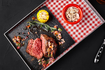 Sliced medium rare grilled Beef steak Ribeye with corn, rosemary, onion and mushroom sauce on a metal tray on a black background
