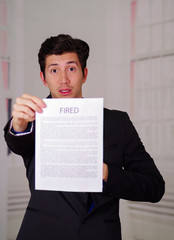 Close up of surprised young businessman wearing a suit and holding a sheet of paper of fired text on it, in a blurred background