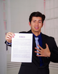 Close up of surprised young businessman wearing a suit and holding a sheet of paper of fired text on it, having a heart attack, in a blurred background