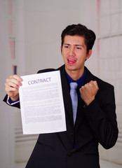 Close up of happy businessman wearing a suit and holding a sheet of paper of contract text on it, in a blurred background