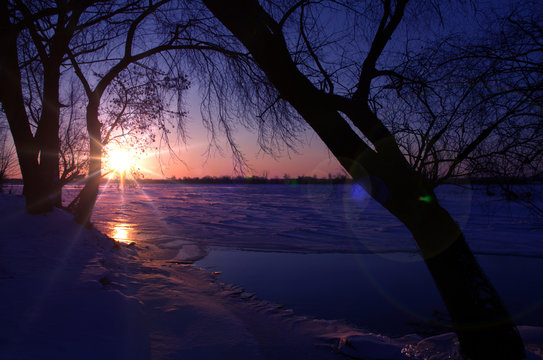 Sunrise between the trees on the banks of the frozen river, which is in the ice of the Bug river in Poland