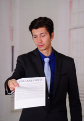 Close up of sad young businessman wearing a suit and holding a sheet of paper getting from the boss, with you are fired text on it, in a blurred background