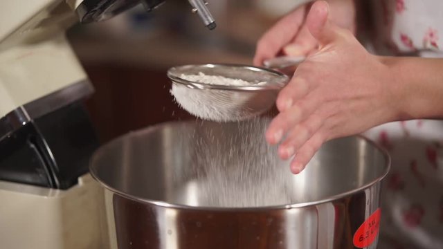Close up shot of a woman strewing a flour in a metal bowl. She is using sieve for better result.