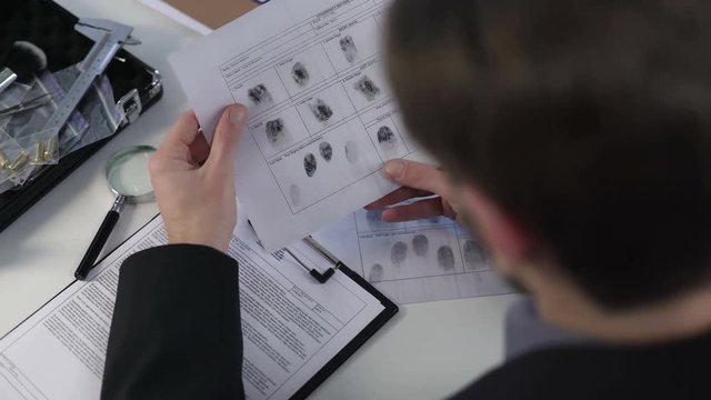 Detective watching fingerprints on paper, using magnifying glass, solving murder