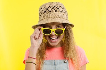 Summer time, a teen girl in a straw hat and sunglasses winking over yellow background.
