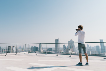 Young white Caucasian man taking cityscape photo on building rooftop on sunny day. Photography hobby, gadget technology, or leisure activity concept. With copy space on blue sky