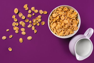 Bowl with corn flakes, jug of milk on purple background, top view